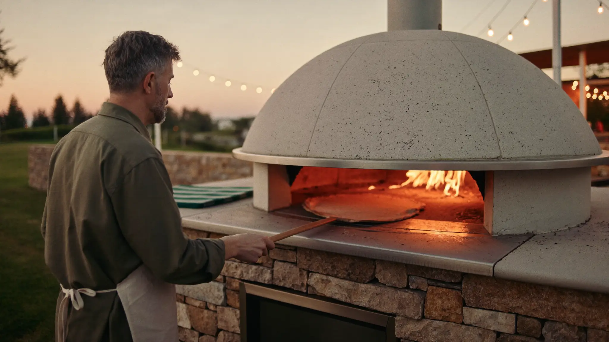 A man at a dome pizza oven on a stone patio at dusk, flames glowing in the oven mouth, peel in hand