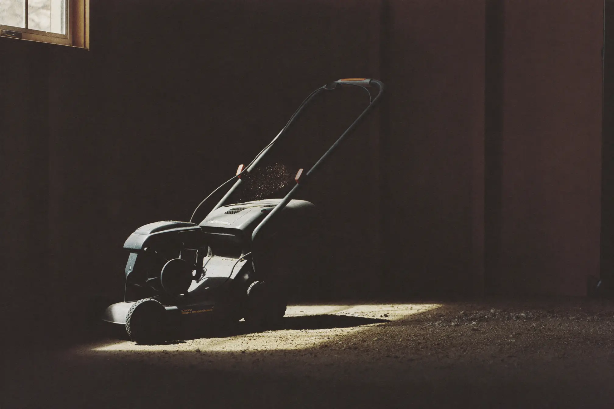 A push lawnmower parked in a dark shed, one blade of window light across it