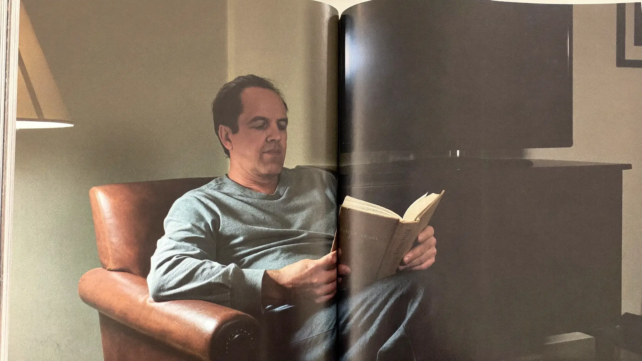 A man reading a hardcover book in an armchair by lamplight, a dark television behind him